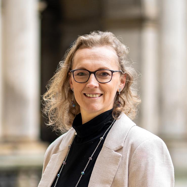 Eine Frau mit Brille und lockigen Haaren trägt ein blaues Sakko und steht vor einer beigen Wand.