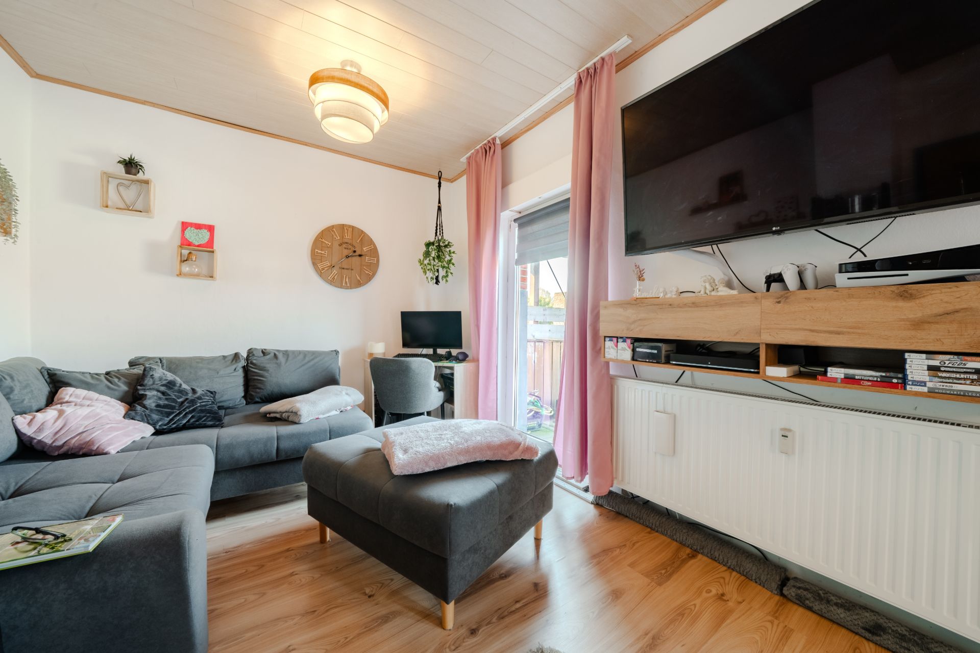 Helles Wohnzimmer mit grauem Sofa, Fernseher an der Wand, Holzregal und rosa Vorhängen.