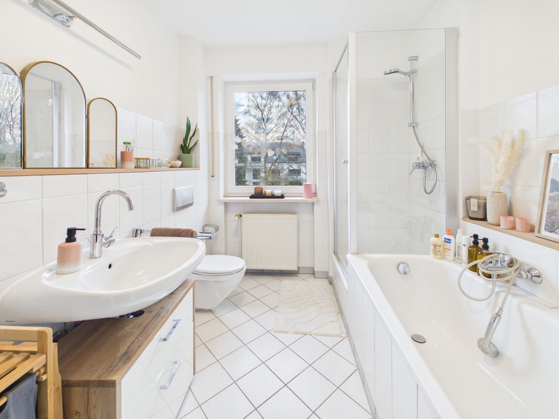 Helles Badezimmer mit Badewanne, Dusche, Waschbecken und Fenster, dekoriert mit Pflanzen und Accessoires.