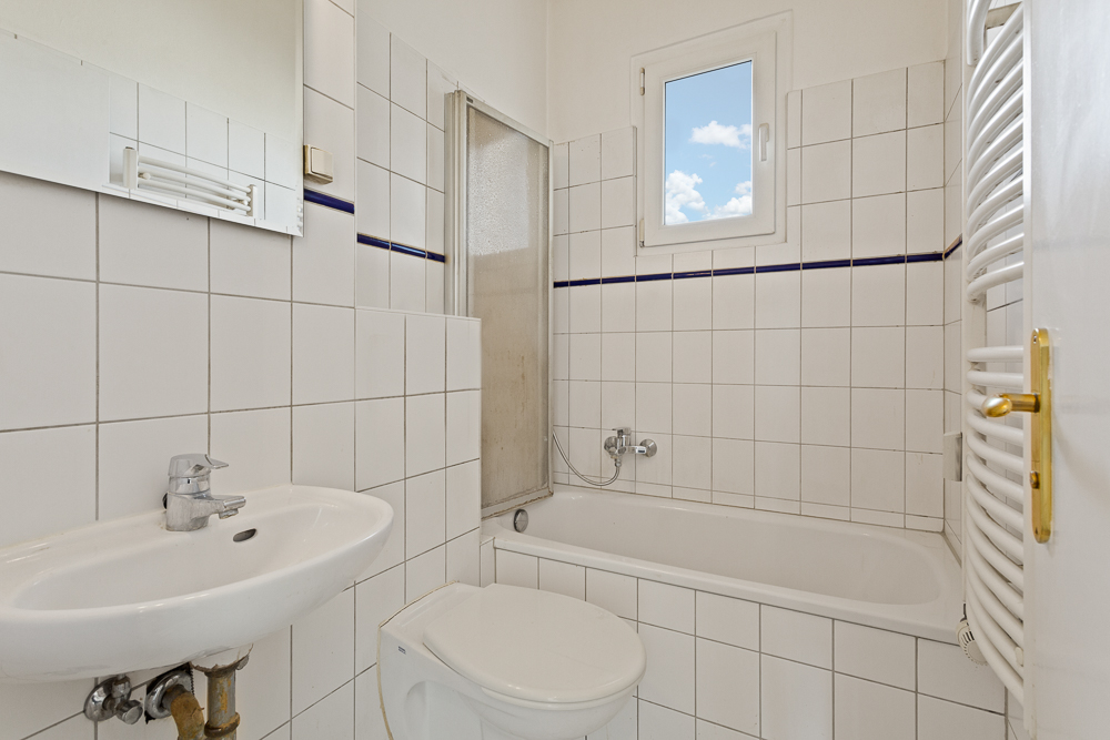 Kleines Badezimmer mit Waschbecken, Toilette, Badewanne, Fenster und weiß gefliesten Wänden.
