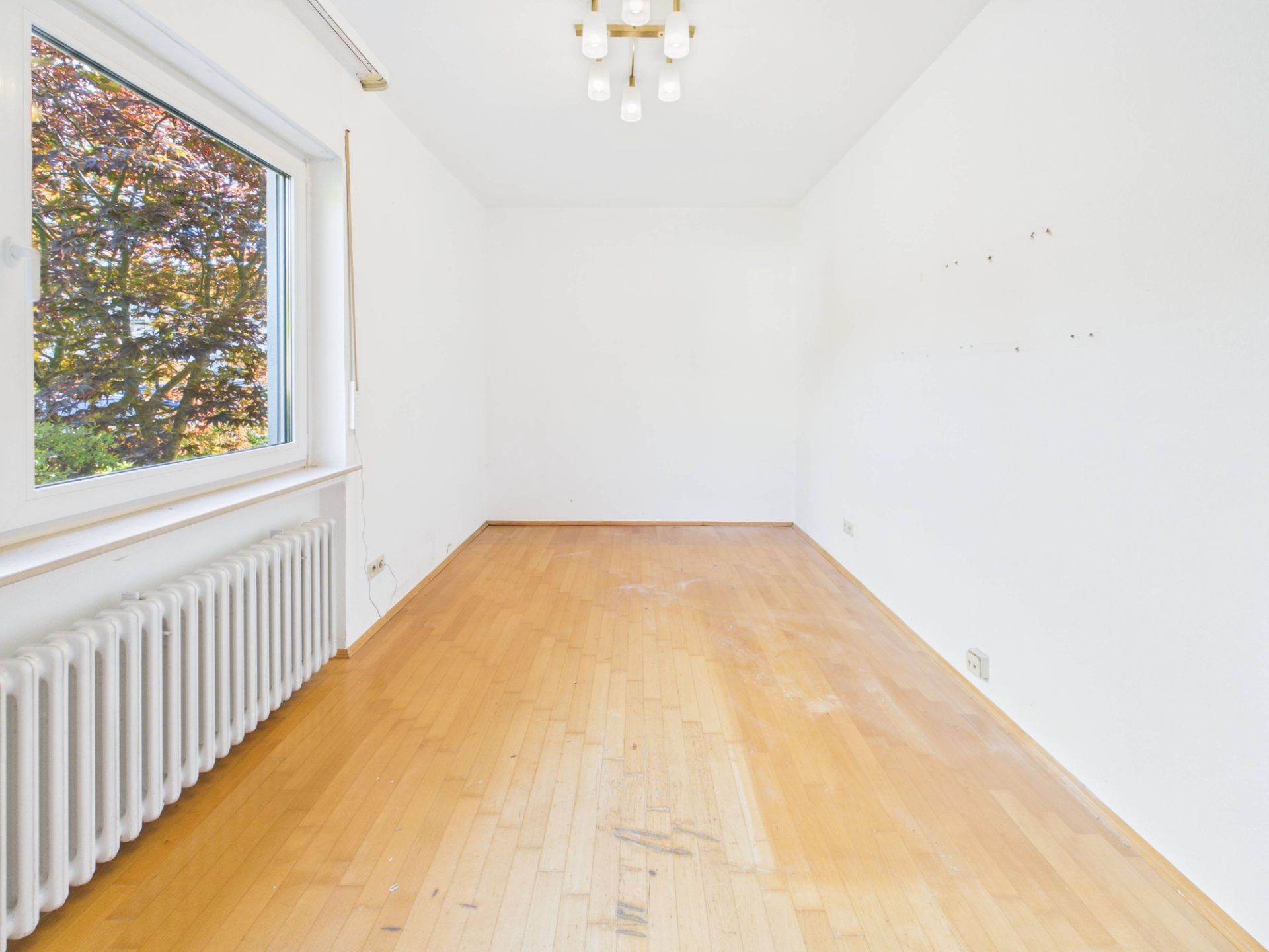 Heller Raum mit Parkettboden, großem Fenster, Heizkörper und weißer Decke mit Lampe.