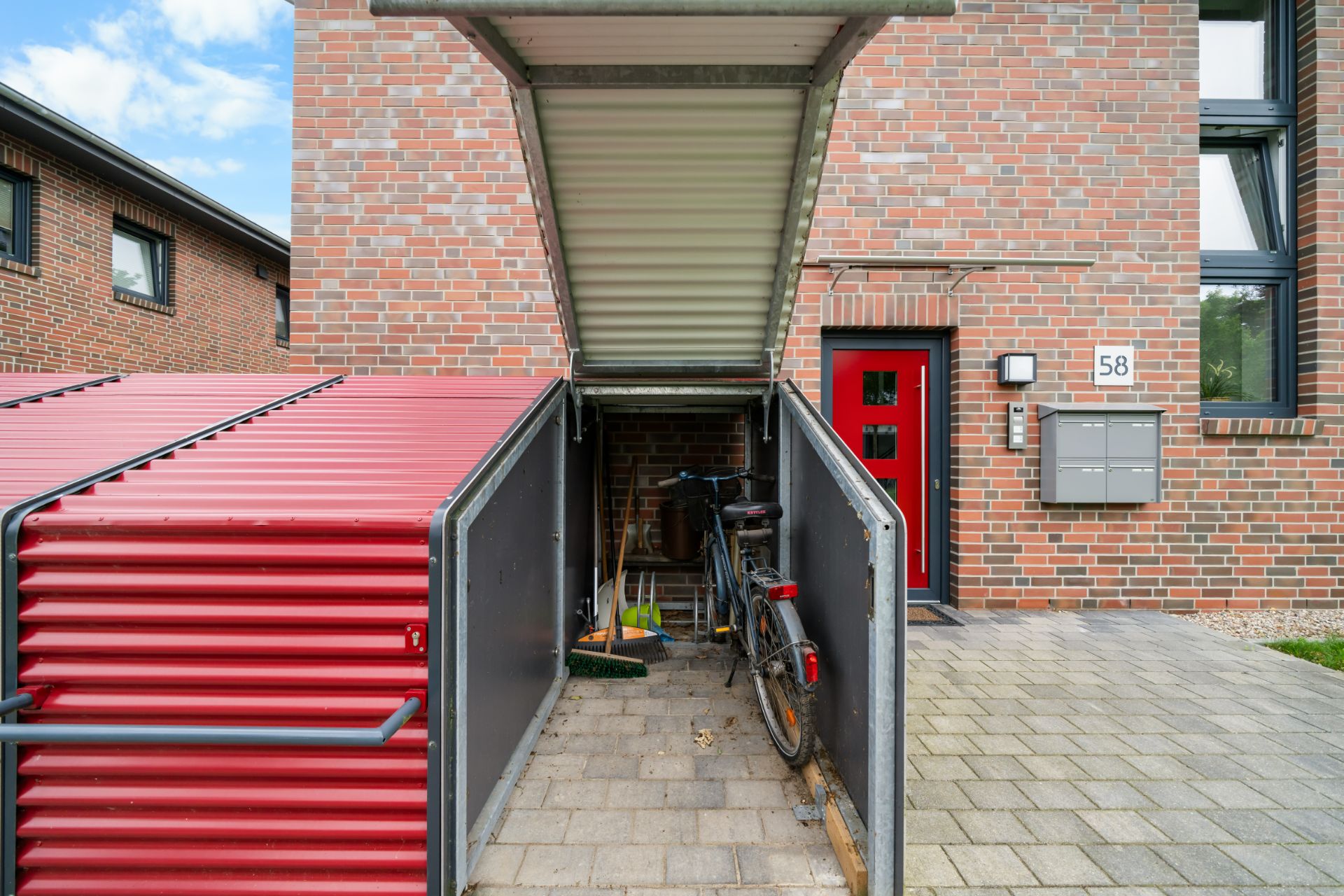 Auf dem Bild ist ein geöffneter Fahrradschuppen mit einem Fahrrad und Gartengeräten vor einem Wohnhaus zu sehen.