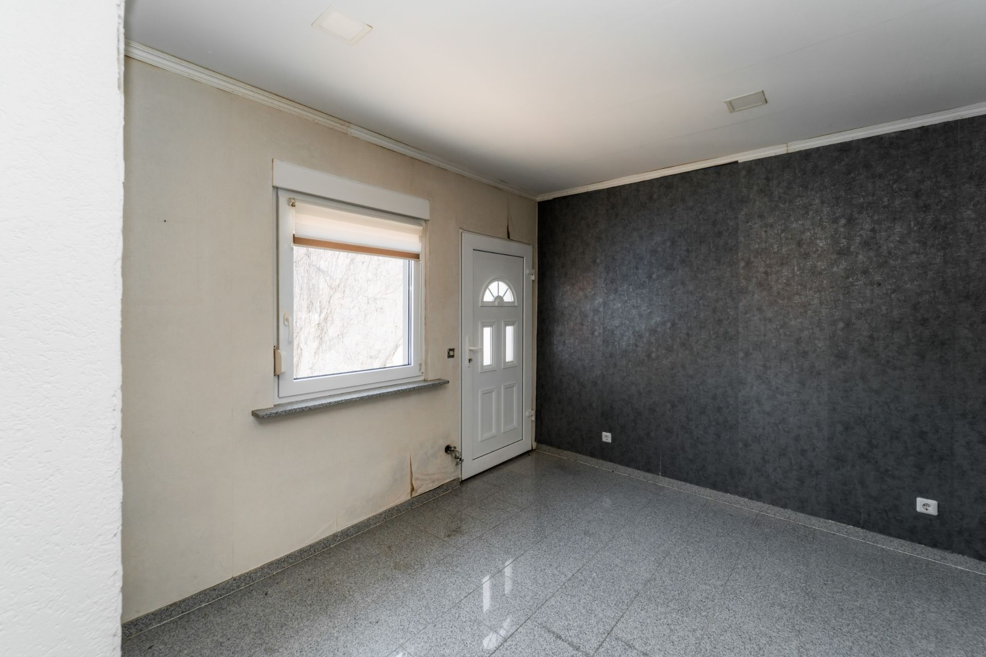 Leerer Raum mit grauem Fliesenboden, Fenster und weißer Eingangstür.