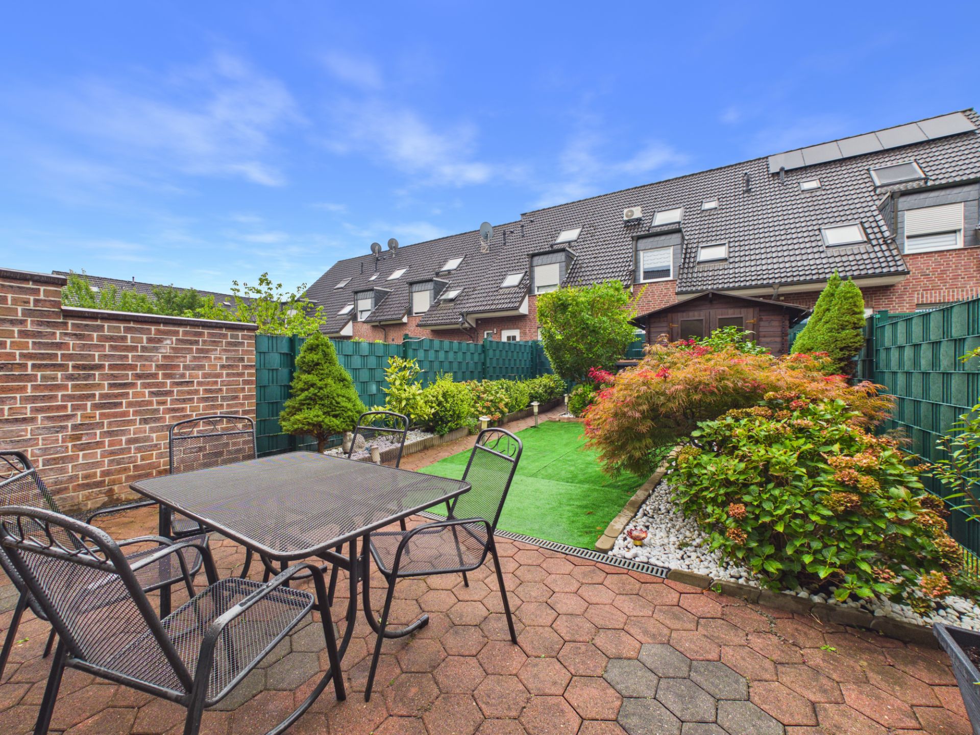 Gepflasterte Terrasse mit Tisch und Stühlen, dahinter ein kleiner Garten mit Sträuchern und Rasen.