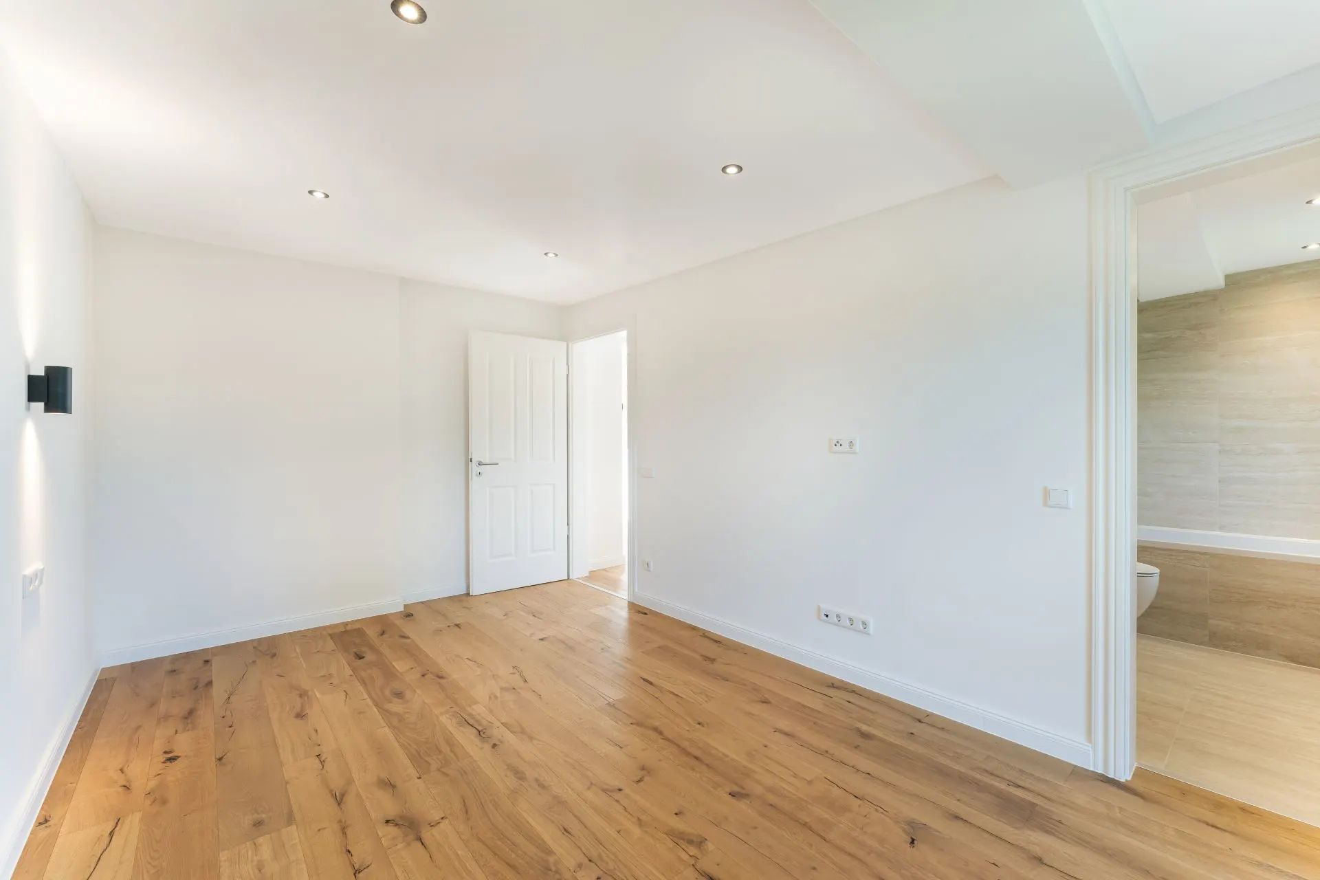 Leerer, heller Raum mit Holzfußboden, weißen Wänden und offener Tür zu einem weiteren Zimmer.
