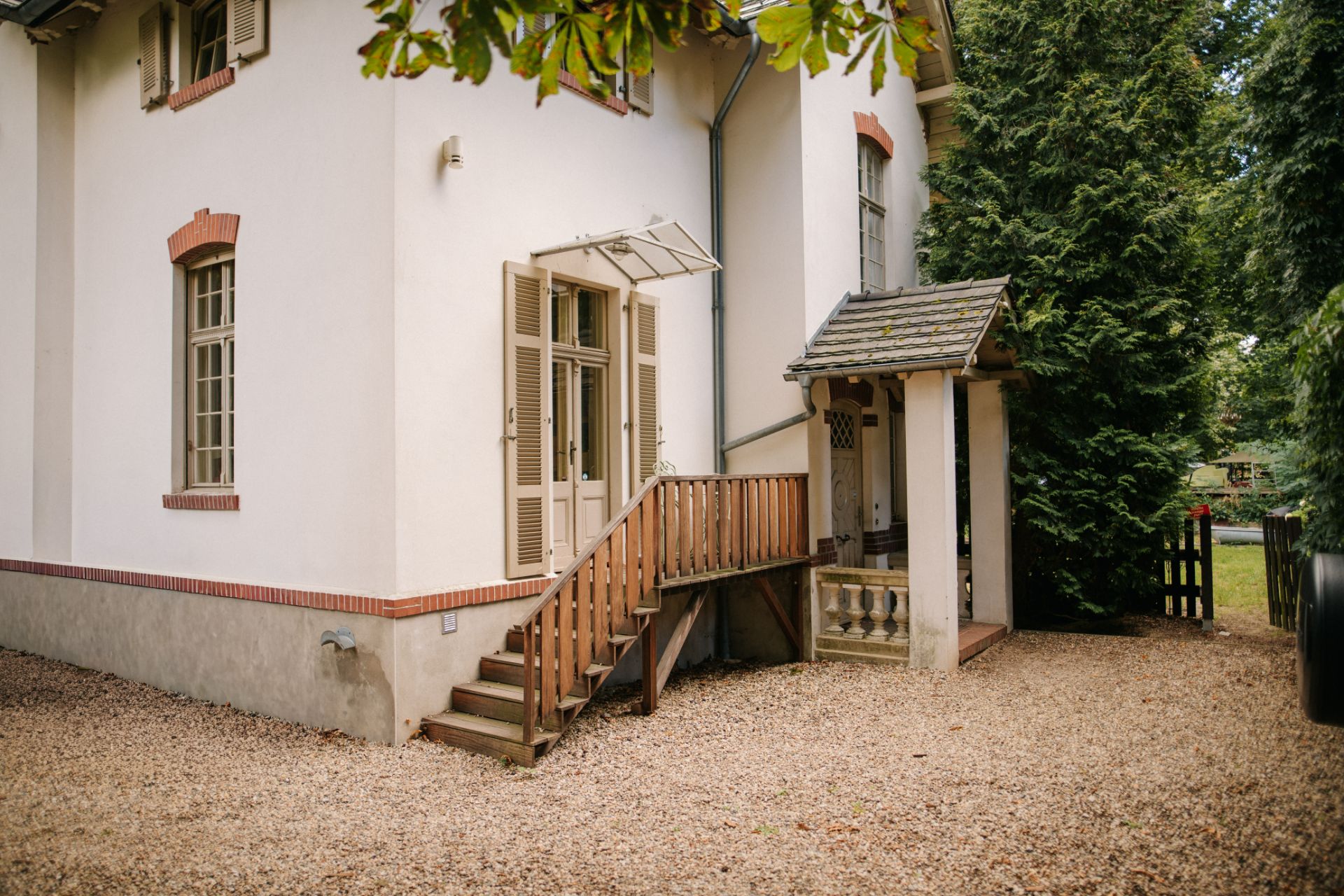Eingang eines weißen Hauses mit Holztreppe, Kiesboden und Fensterläden.