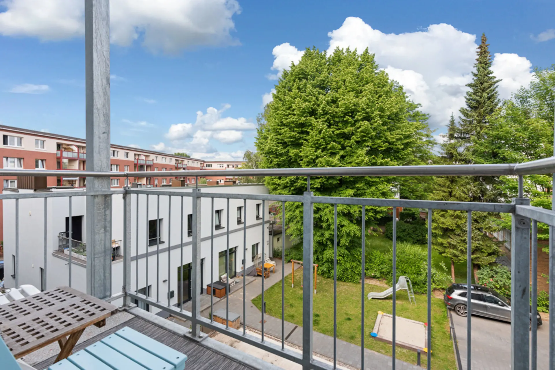 Blick von einem Balkon auf einen grünen Innenhof mit Spielplatz, Sandkasten und Bäumen.
