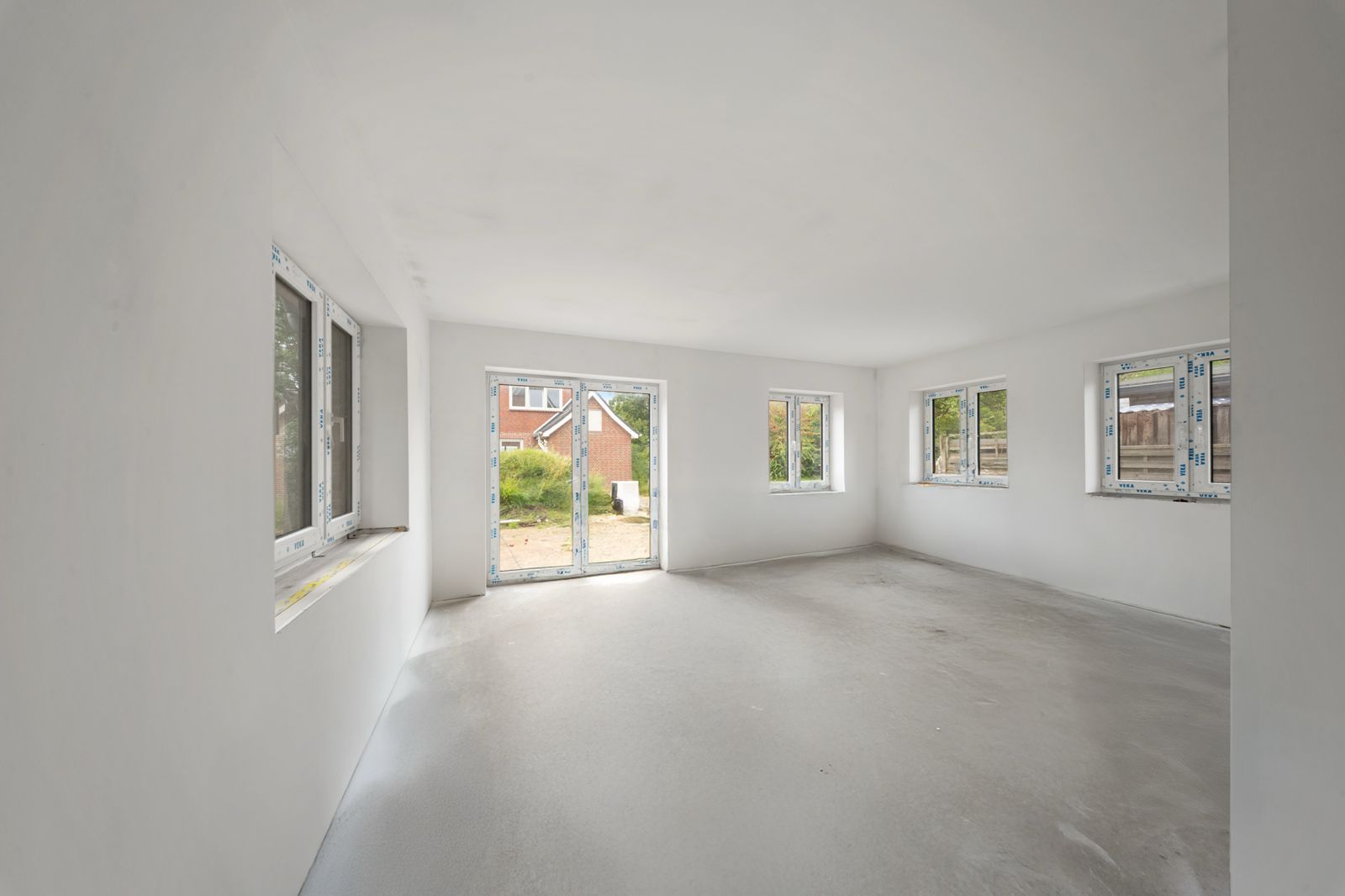 Heller, leerer Raum mit mehreren Fenstern und Glastür, Blick nach draußen auf einen Garten.