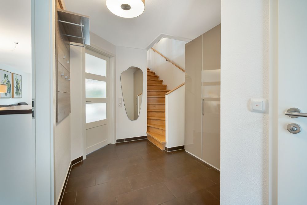 Heller Flur mit Holztreppe, Spiegel, Einbauschränken und braunem Fliesenboden.