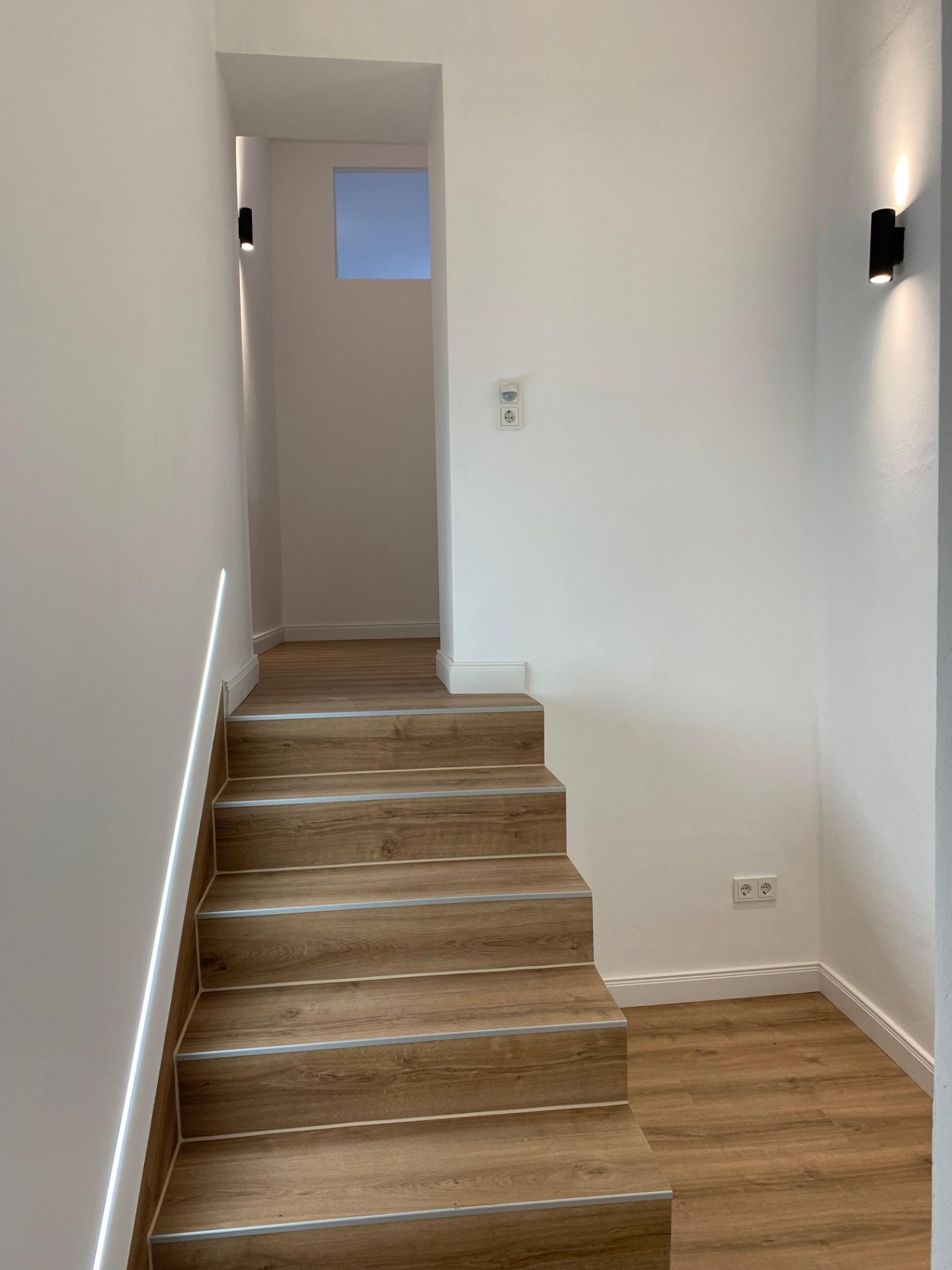 Treppe mit Holzstufen in einem hellen Flur mit weißen Wänden und Wandlampen.