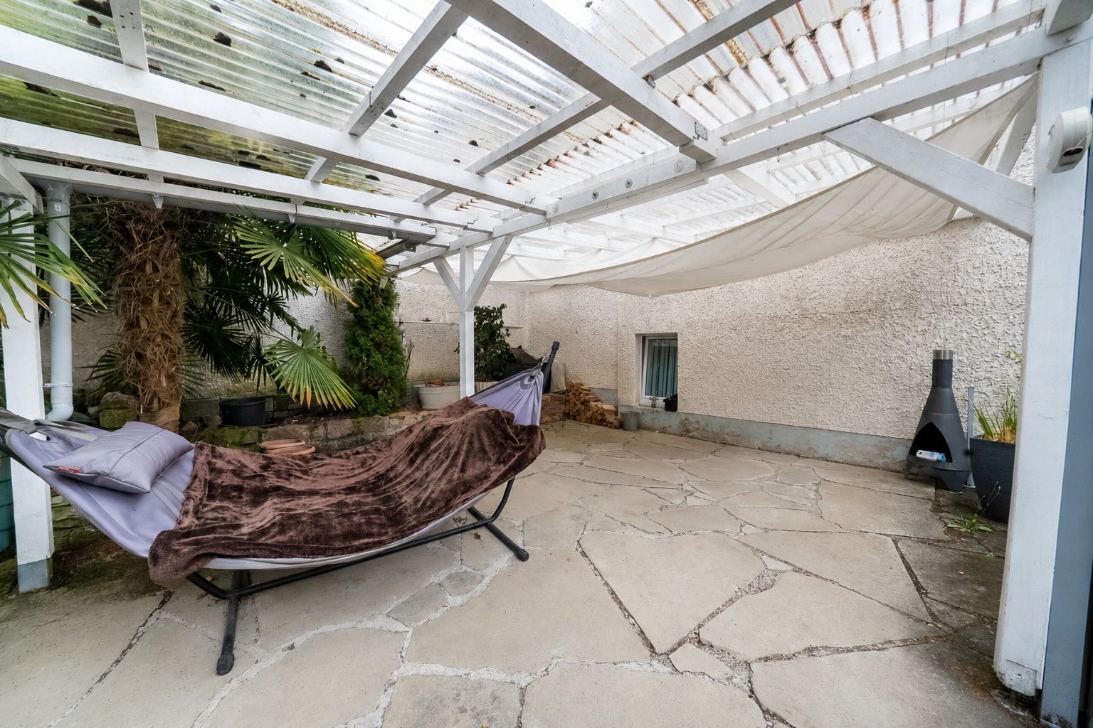 Überdachte Terrasse mit Hängematte, Decke, Pflanzen und Außenkamin auf Steinboden.