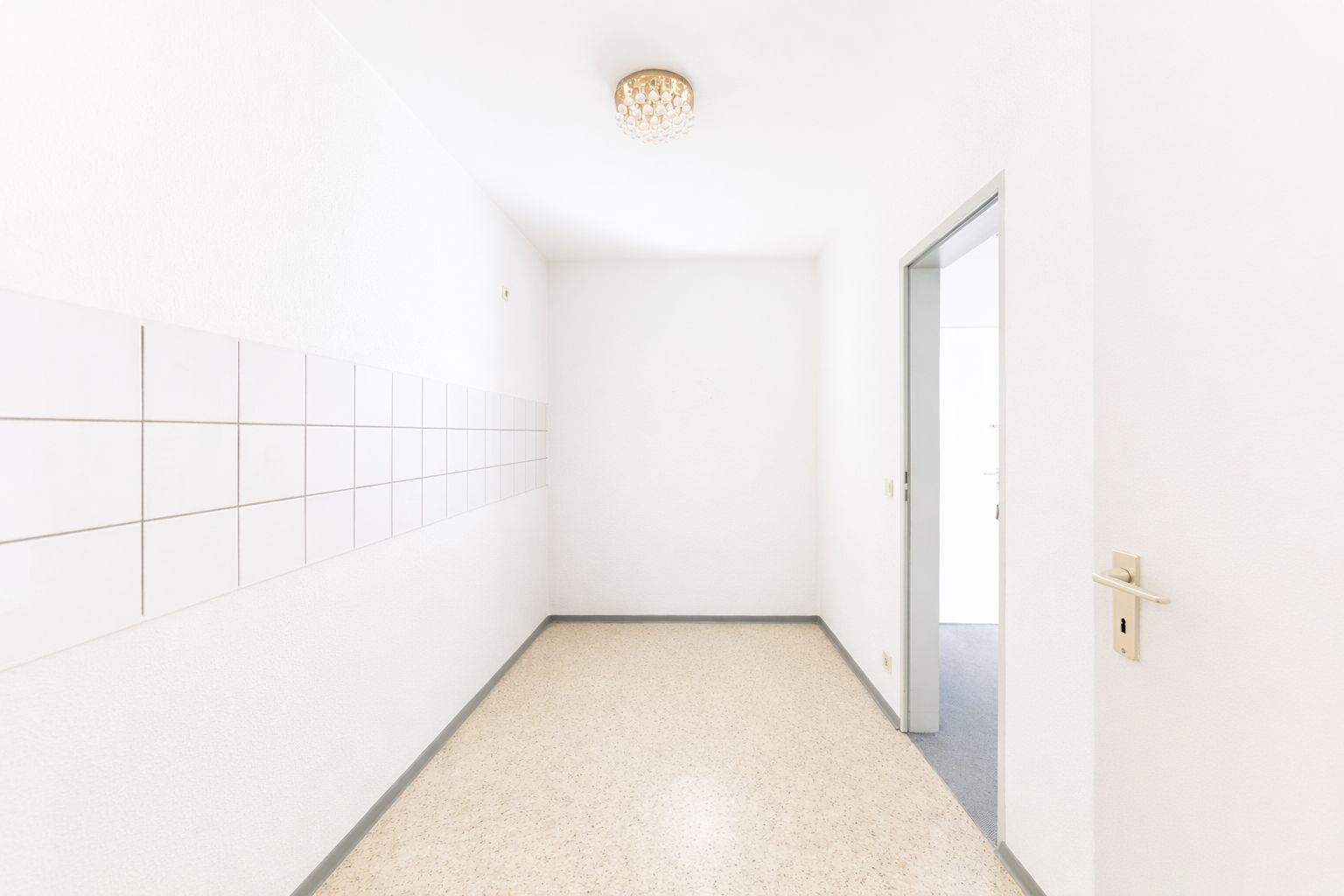 Heller leerer Raum mit Fliesen an einer Wand, Türöffnung rechts, Deckenlampe.