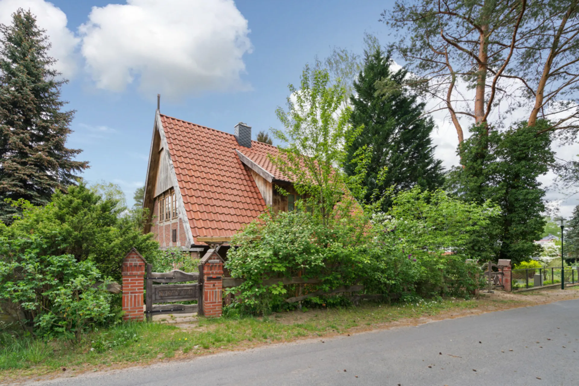 Haus mit rotem Dach hinter Holzzaun und viel grünem Bewuchs an einer ruhigen Straße.