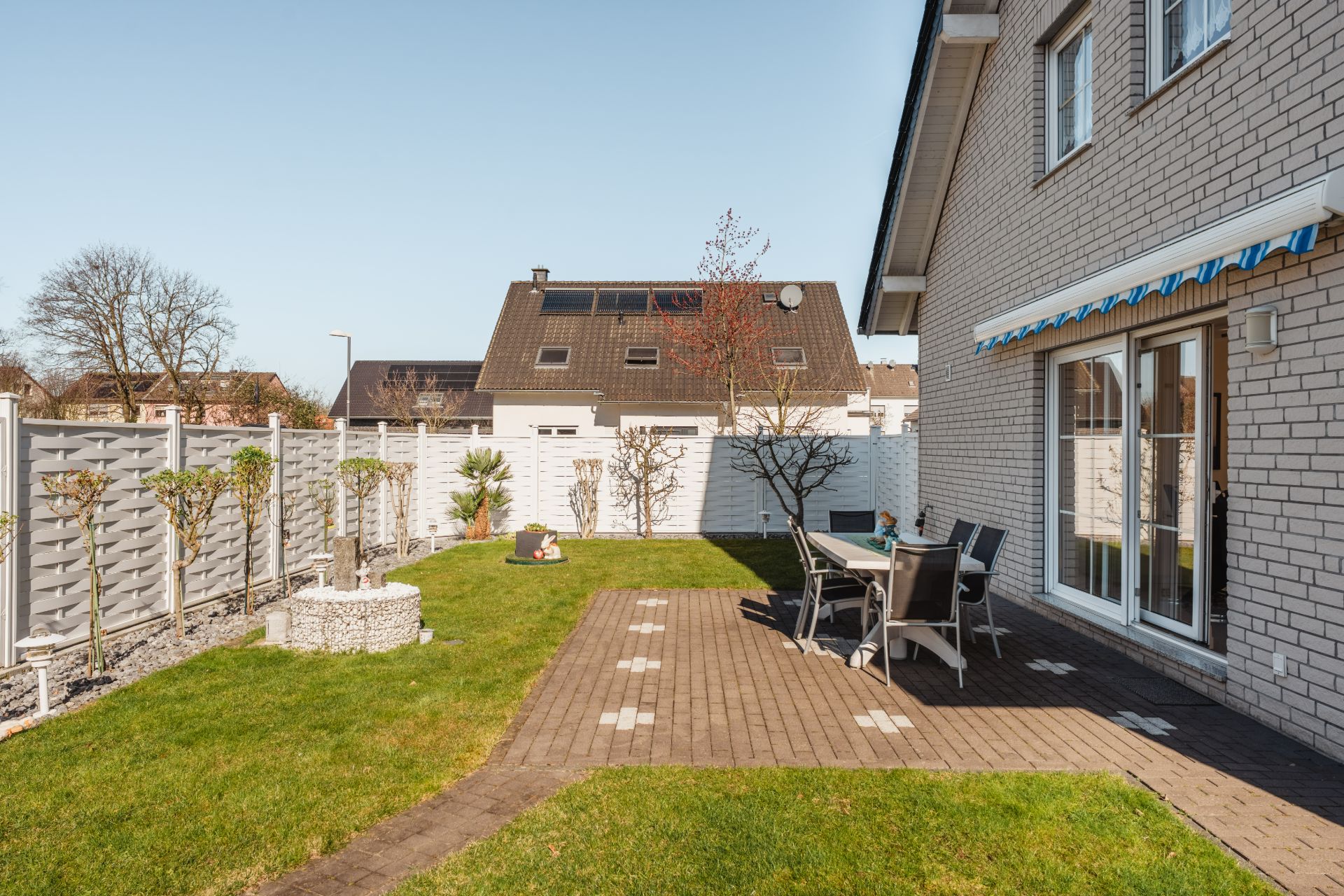 Garten mit Rasen, Terrasse, Esstisch und Stühlen neben einem Haus mit weißen Ziegelwänden.