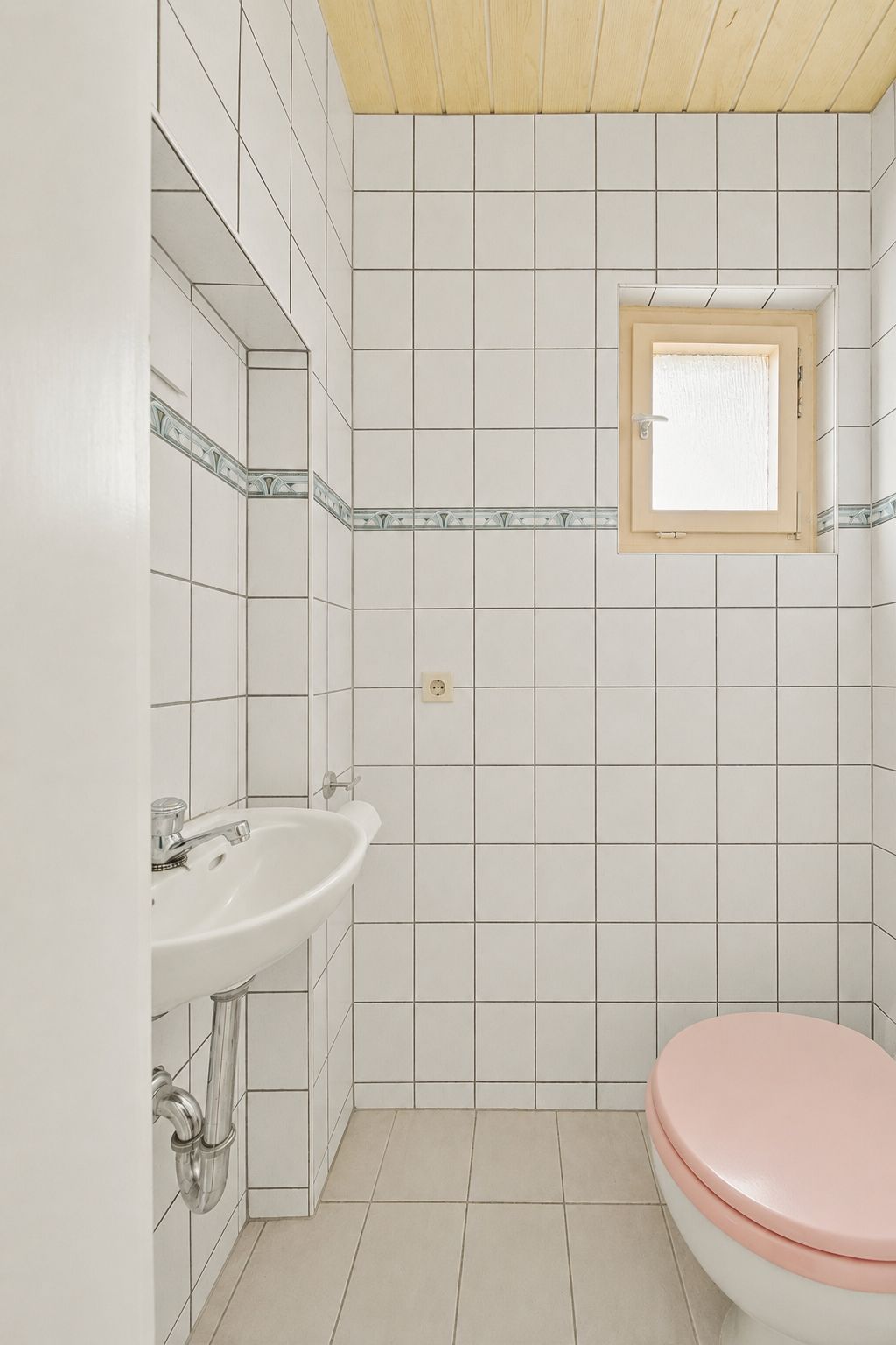Kleines Badezimmer mit weiß gefliesten Wänden, Waschbecken, Fenster und Toilette mit rosa Deckel.