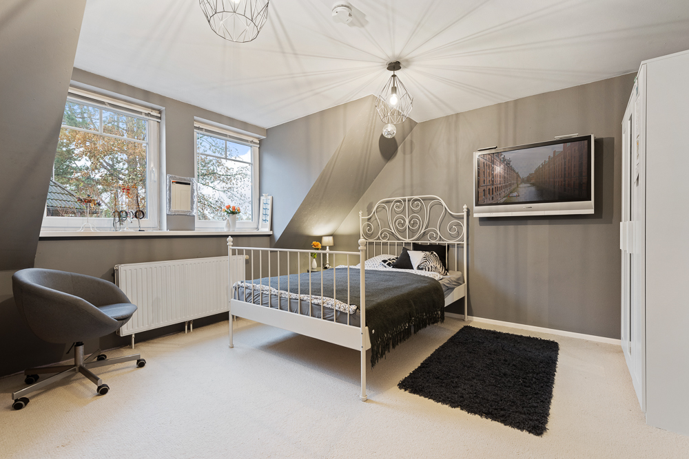 Helles Schlafzimmer mit weißem Bett, grauen Wänden, Teppich, Schreibtischstuhl und zwei Fenstern.
