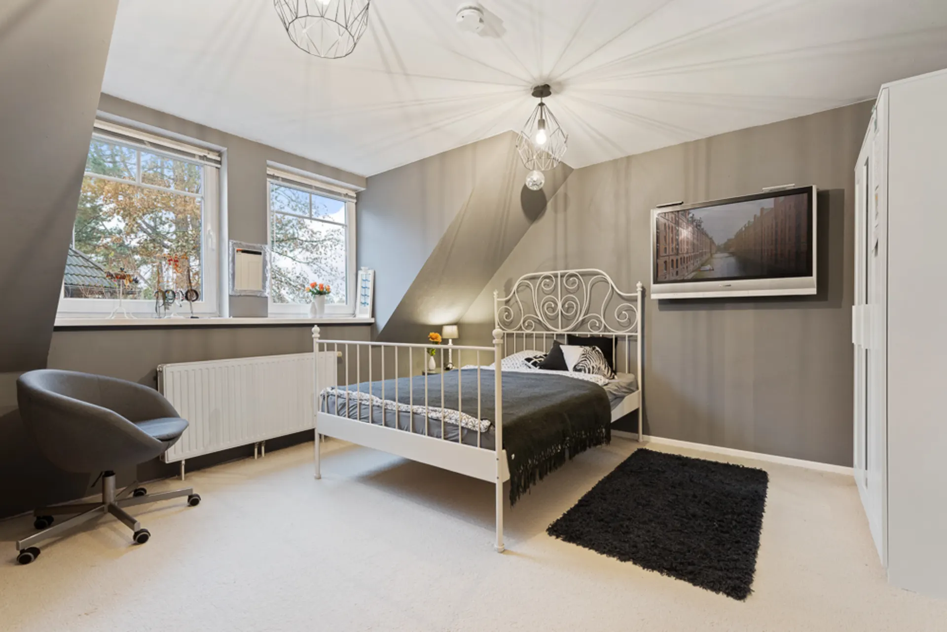 Helles Schlafzimmer mit weißem Bett, grauen Wänden, Teppich, Schreibtischstuhl und zwei Fenstern.