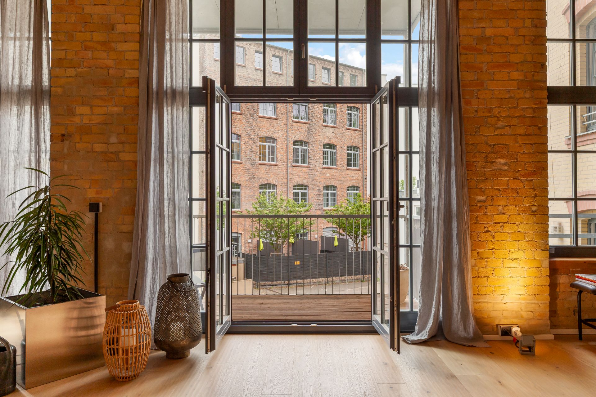 Große Glas-Doppeltüren führen auf einen Balkon, dahinter ein Backsteingebäude und grüne Pflanzen.