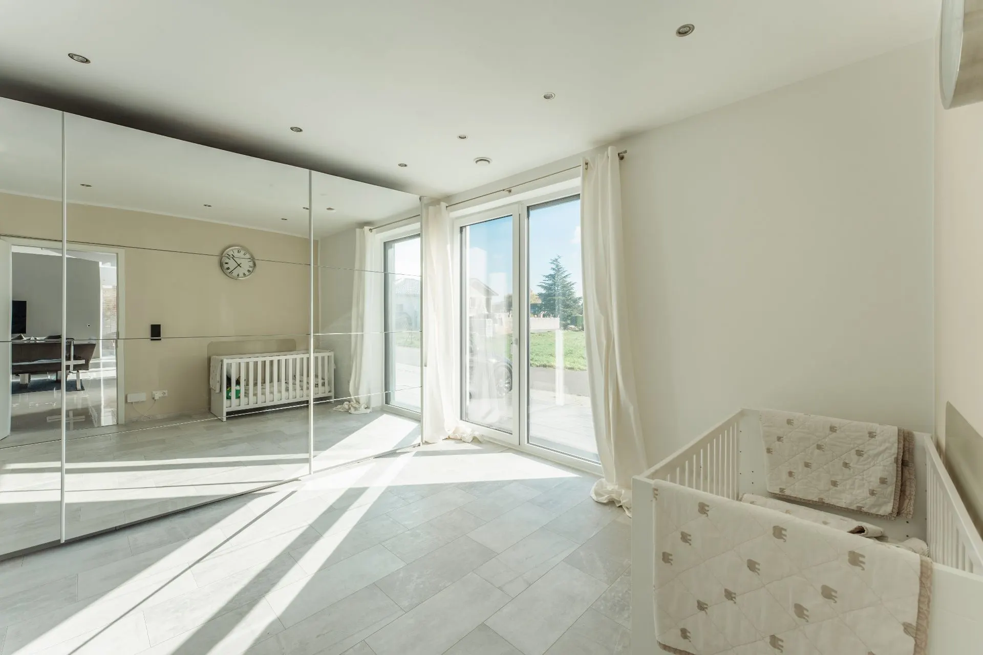 Helles Zimmer mit Babybett, Spiegelwand und großen Fenstern mit Blick nach draußen.