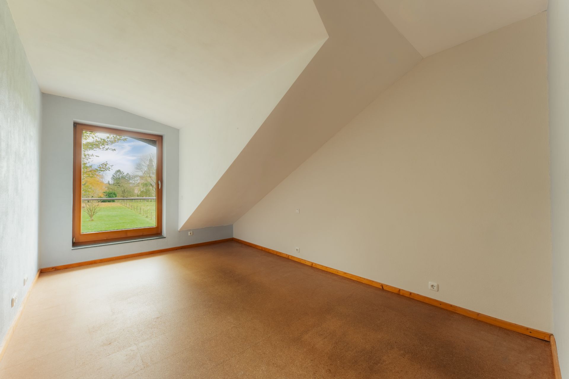 Leeres Zimmer mit schräger Decke, großem Fenster und Blick auf einen Garten.