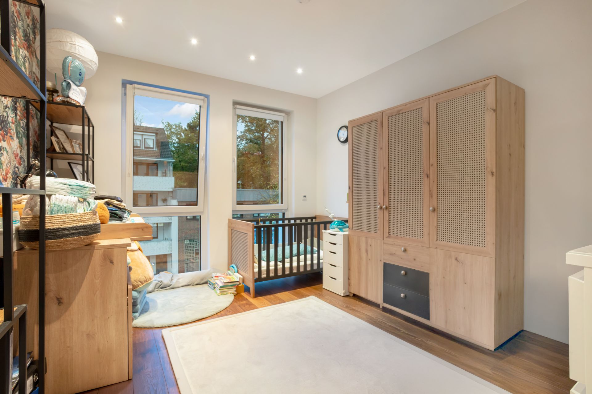 Helles Kinderzimmer mit Babybett, Teppich, Schrank und Wickeltisch, zwei Fenster mit Ausblick.