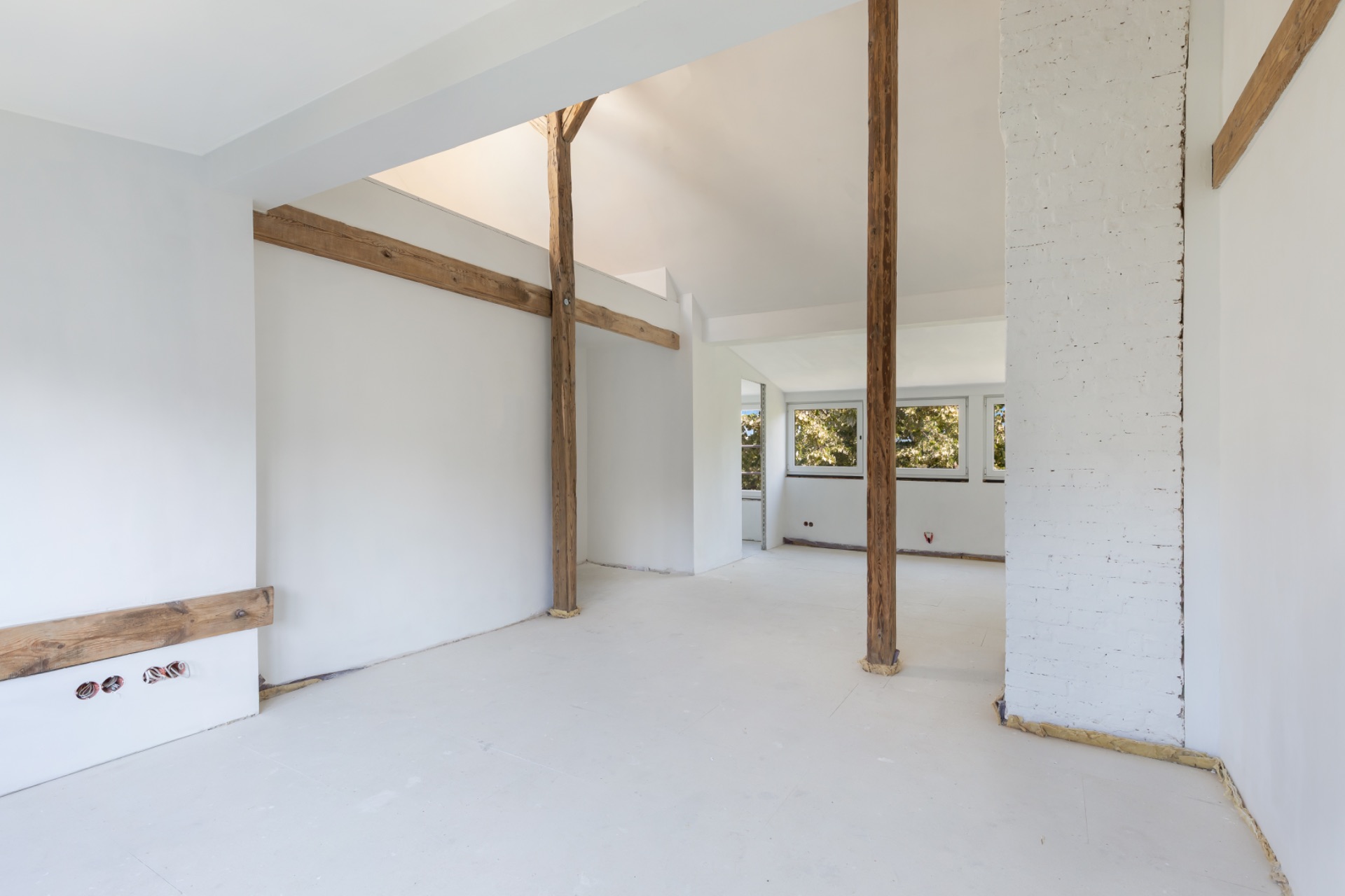 Leerer, heller Raum mit weißen Wänden und Holzbalken, mehrere Fenster im Hintergrund.