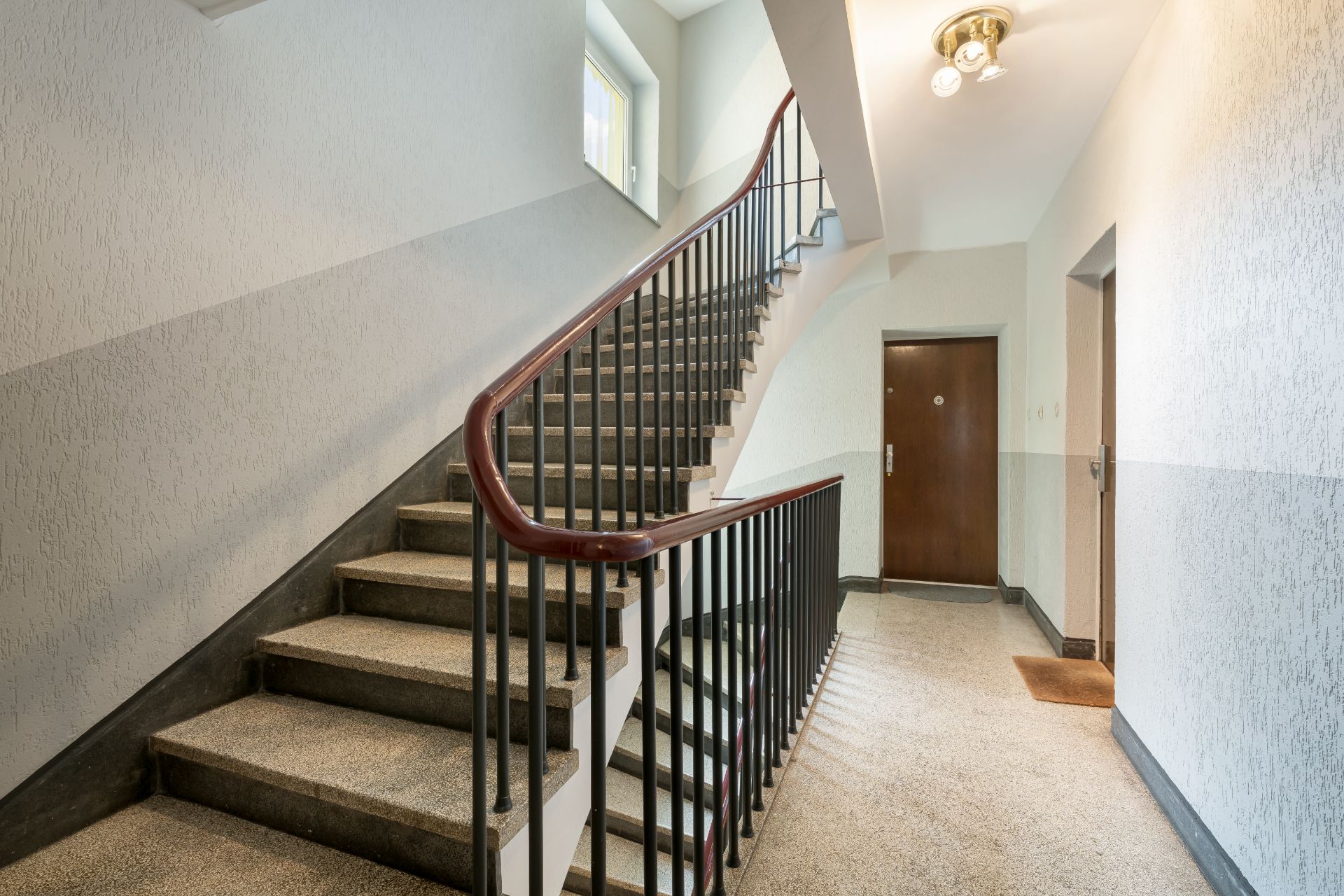Helles Treppenhaus mit Metallgeländer und Wohnungstür, Fenster oben, Deckenlampe.