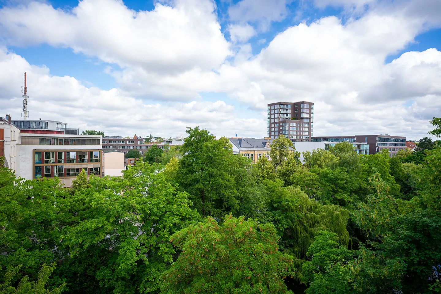 Blick über viele grüne Bäume auf mehrere moderne Wohngebäude unter blauem Himmel mit Wolken.