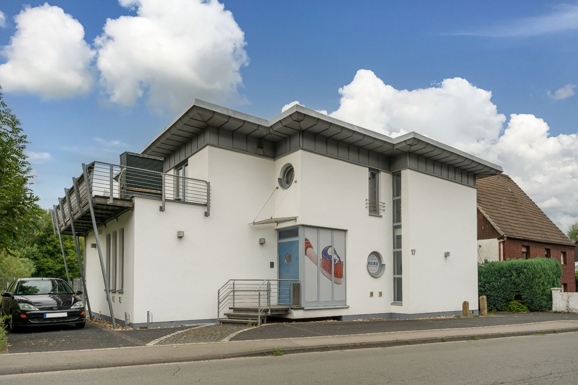 Modernes, weißes Wohnhaus mit Balkon, Treppe am Eingang und geparktem Auto daneben.