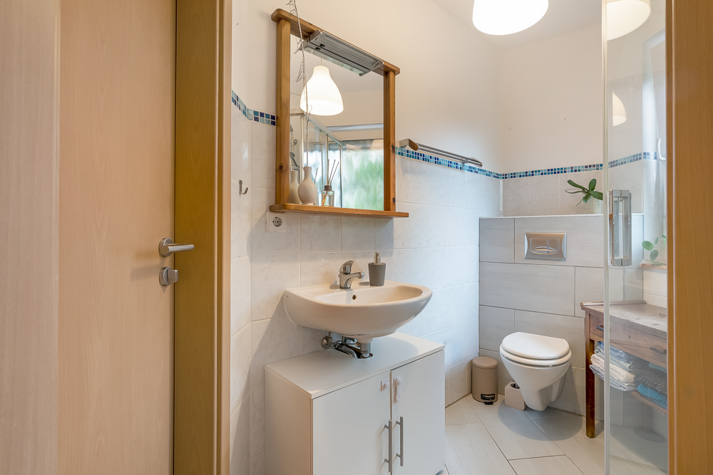 Kleines Badezimmer mit Waschbecken, Spiegel, WC und Fenster. Handtücher liegen auf einem Holztisch.