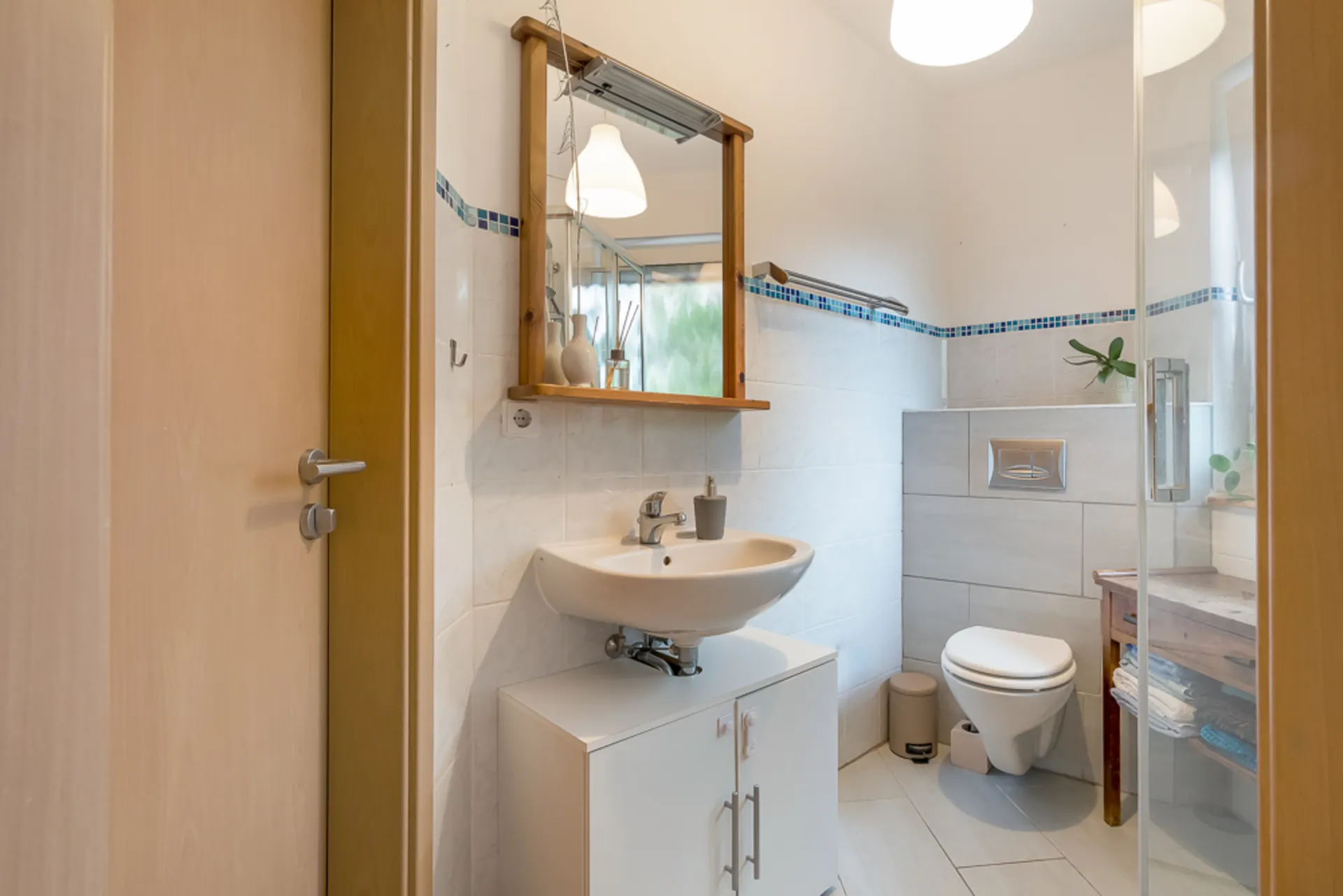 Kleines Badezimmer mit Waschbecken, Spiegel, WC und Fenster. Handtücher liegen auf einem Holztisch.