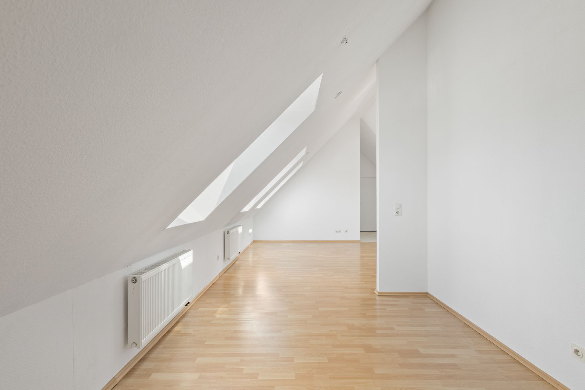 Heller Raum mit Dachschrägen, großen Fenstern und hellem Holzboden.
