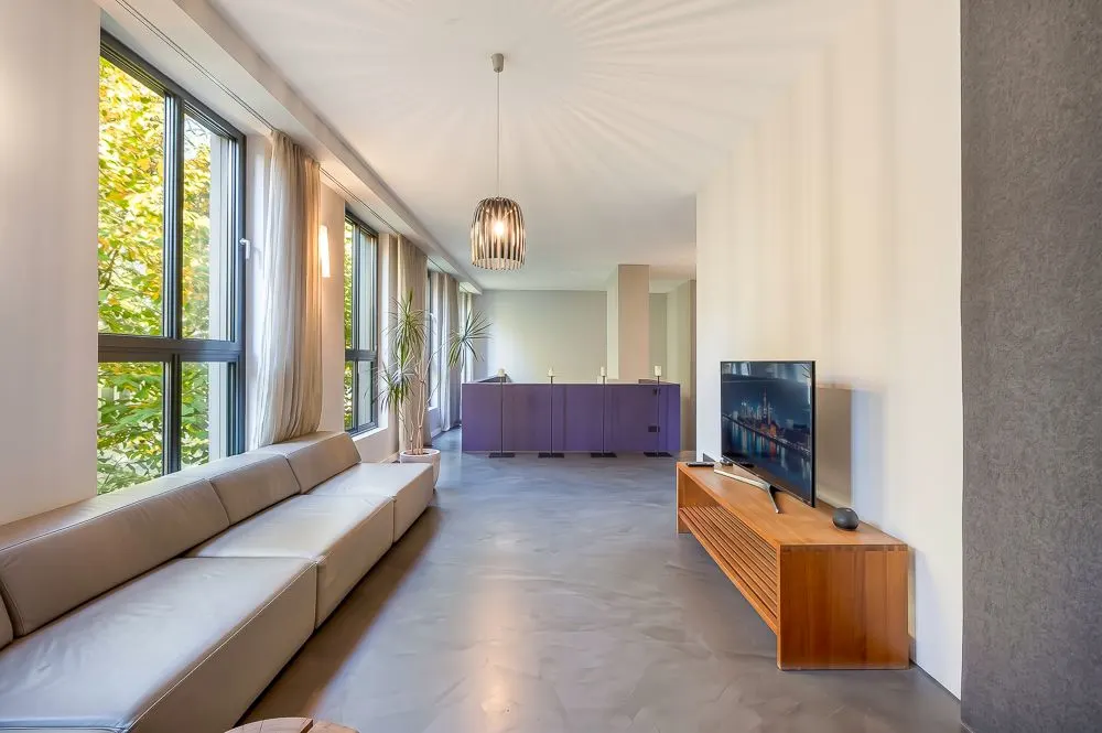 Helles Wohnzimmer mit großer Fensterfront, grauem Sofa, TV auf Holzschrank und lila Sideboard.