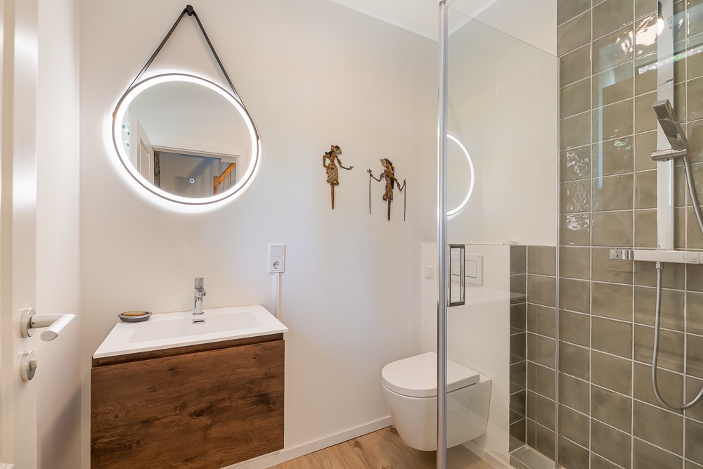 Modernes Badezimmer mit rundem Spiegel, Waschbecken, WC und Dusche mit grünen Fliesen.