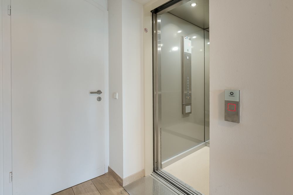 Geöffneter Aufzug neben einer weißen Zimmertür in einem Flur mit Holzboden.