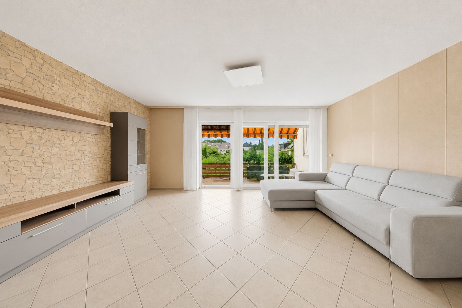 Helles Wohnzimmer mit Ecksofa, Regalen, Steinwand und großen Fenstern mit Blick auf Terrasse.
