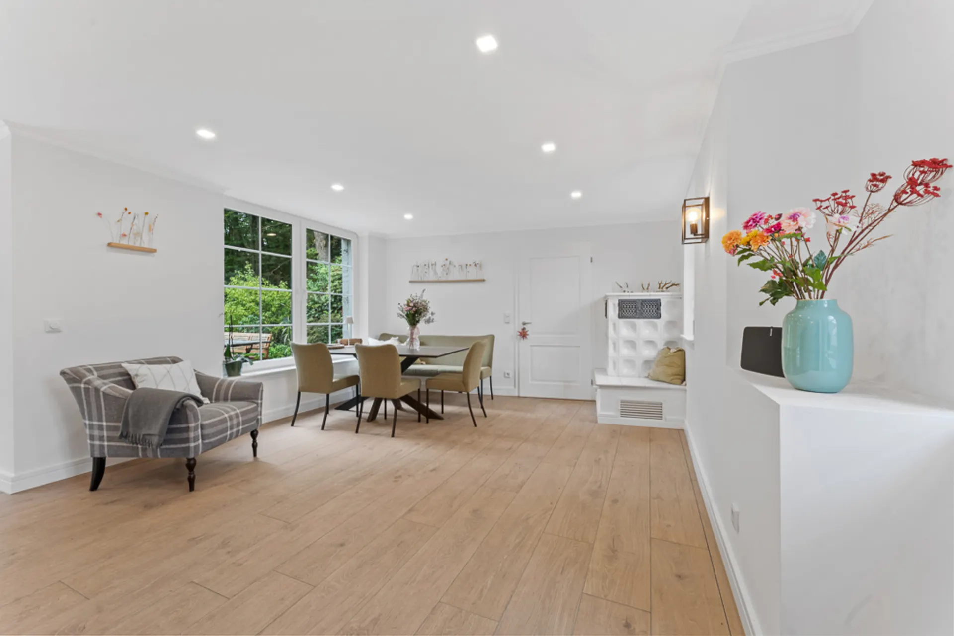 Helles Wohnzimmer mit Esstisch, Sesseln, großen Fenstern und Dekoration mit Blumen.