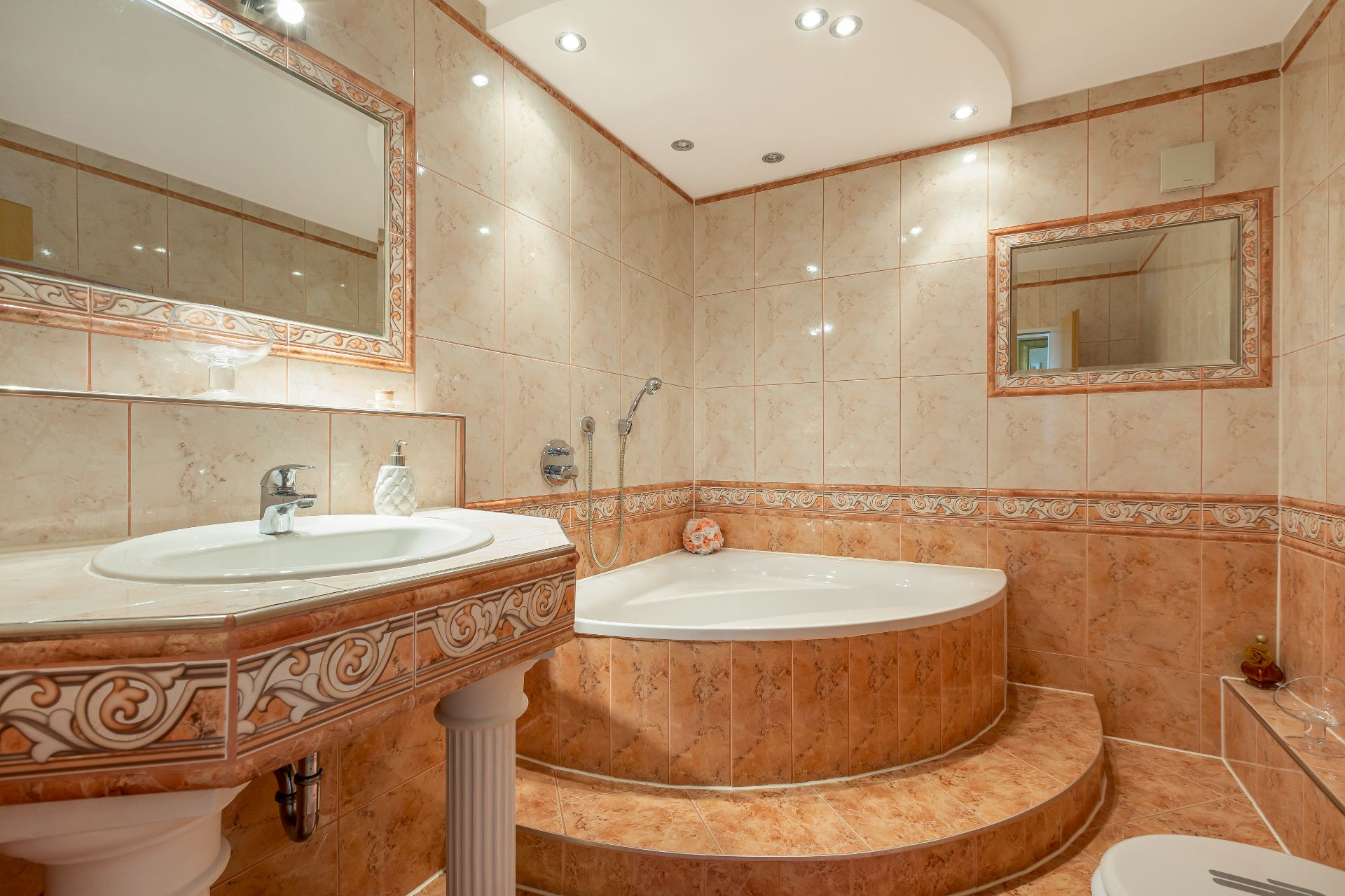 Badezimmer mit Eckbadewanne, großem Spiegel und Waschbecken, alles mit beigen Fliesen.
