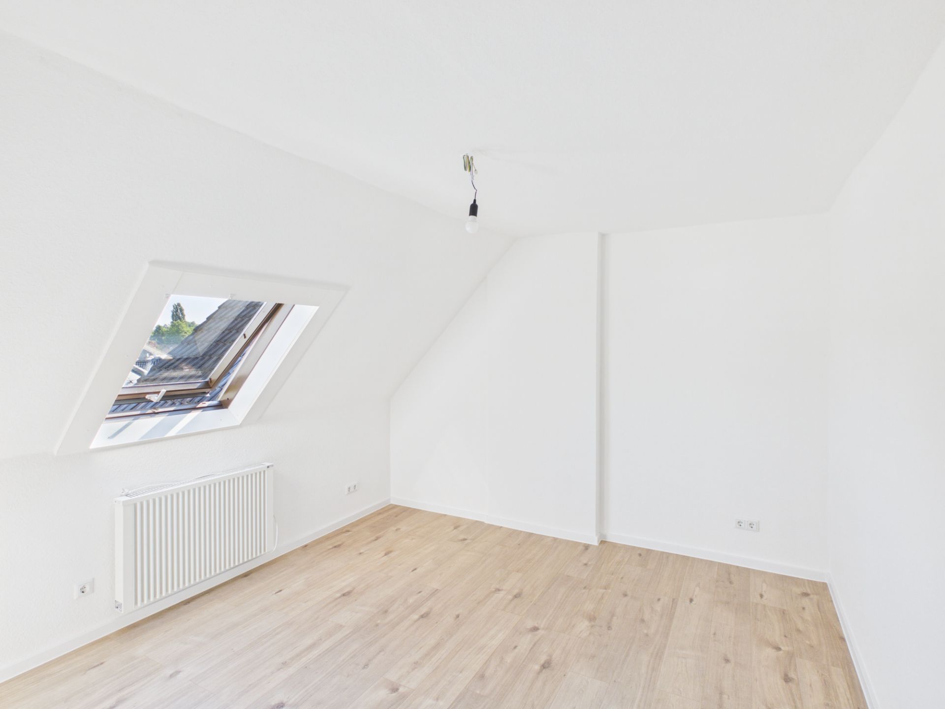 Helles, leeres Zimmer mit Dachschräge, Holzfußboden und großem Fenster mit Heizkörper darunter.