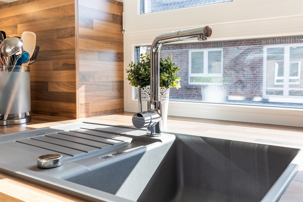 Moderne Küchenspüle mit Wasserhahn, Pflanze und Kochutensilien auf Holzarbeitsplatte vor Fenster.