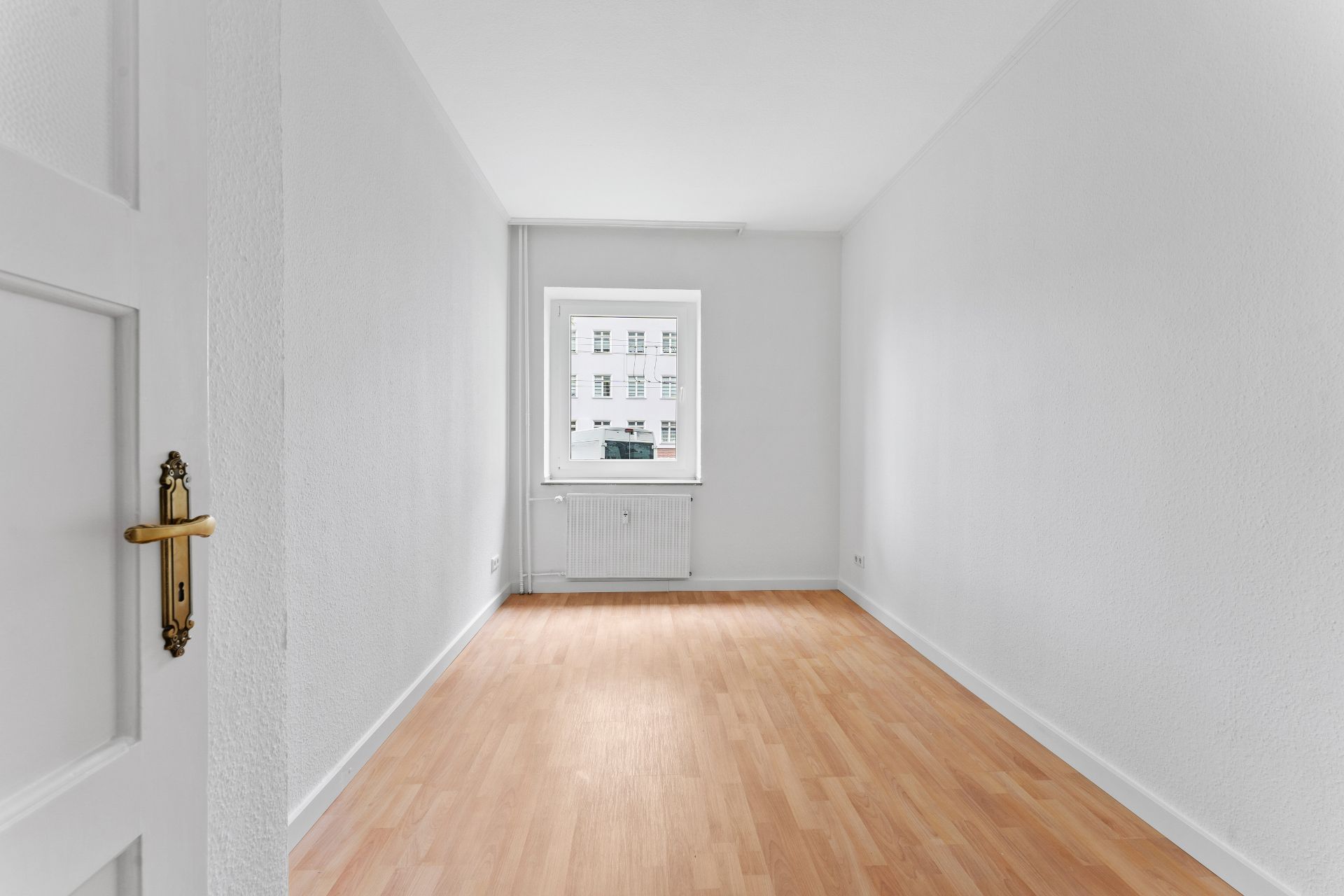 Leeres, helles Zimmer mit Holzfußboden und weißem Fenster an der Stirnseite.