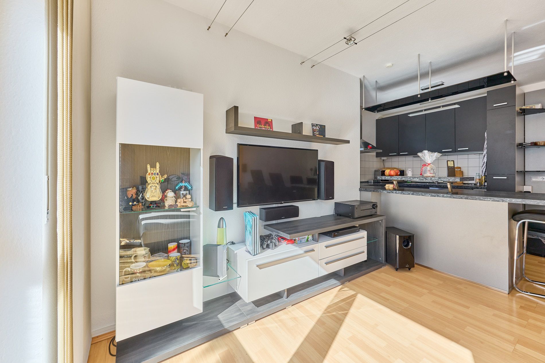 Modernes Wohnzimmer mit TV-Wand, Vitrine und offener Küche mit Barhockern.