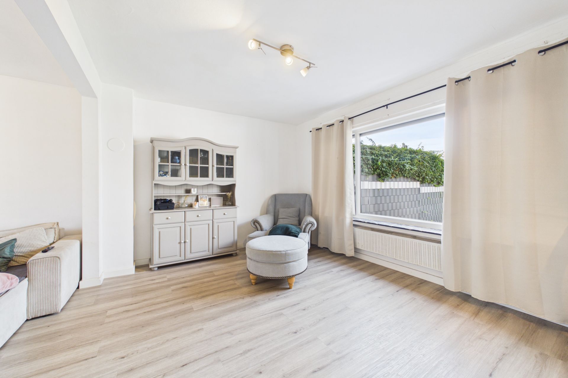 Helles Wohnzimmer mit Sessel, Hocker, Vitrine und großem Fenster mit Vorhängen.