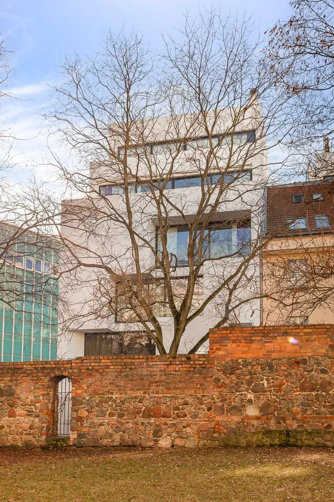 Modernes weißes Wohngebäude hinter einer Steinmauer und einem kahlen Baum.