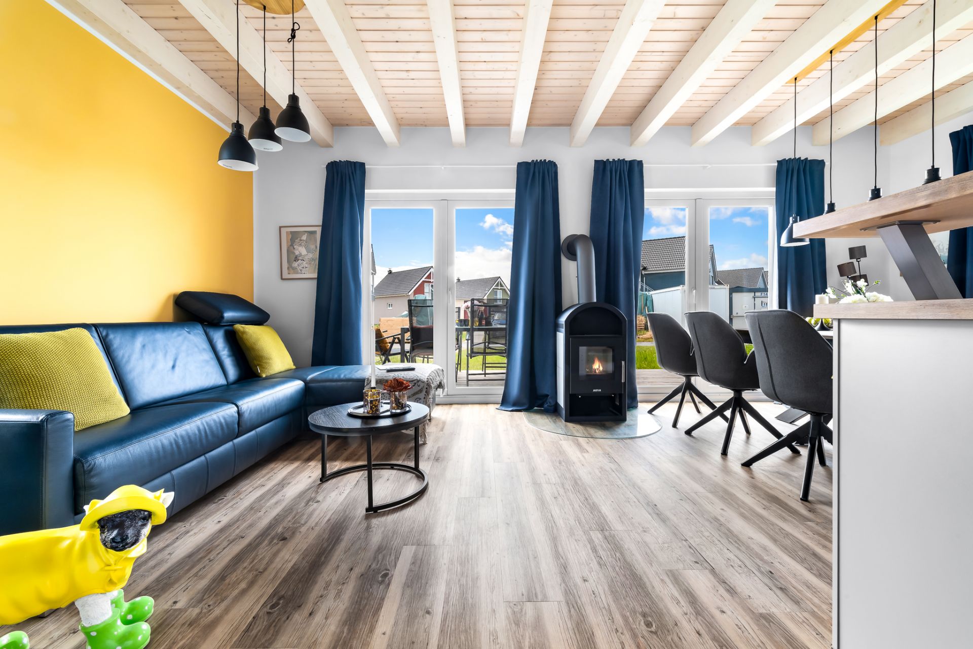 Modernes Wohnzimmer mit gelbem Sofa, Holzboden, Kaminofen und großen Fenstern mit blauen Vorhängen.
