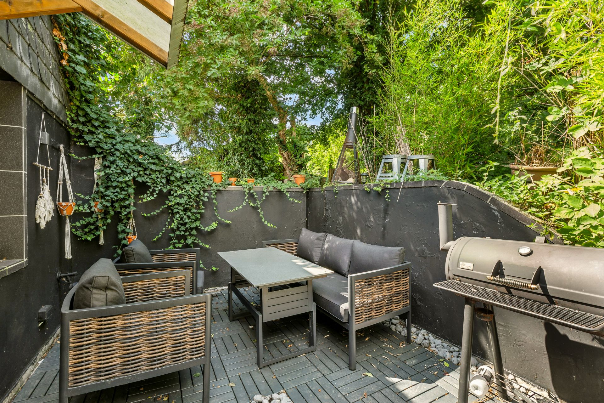 Kleiner Gartenbereich mit Sitzgruppe, Tisch und Grill, umgeben von grünen Pflanzen.