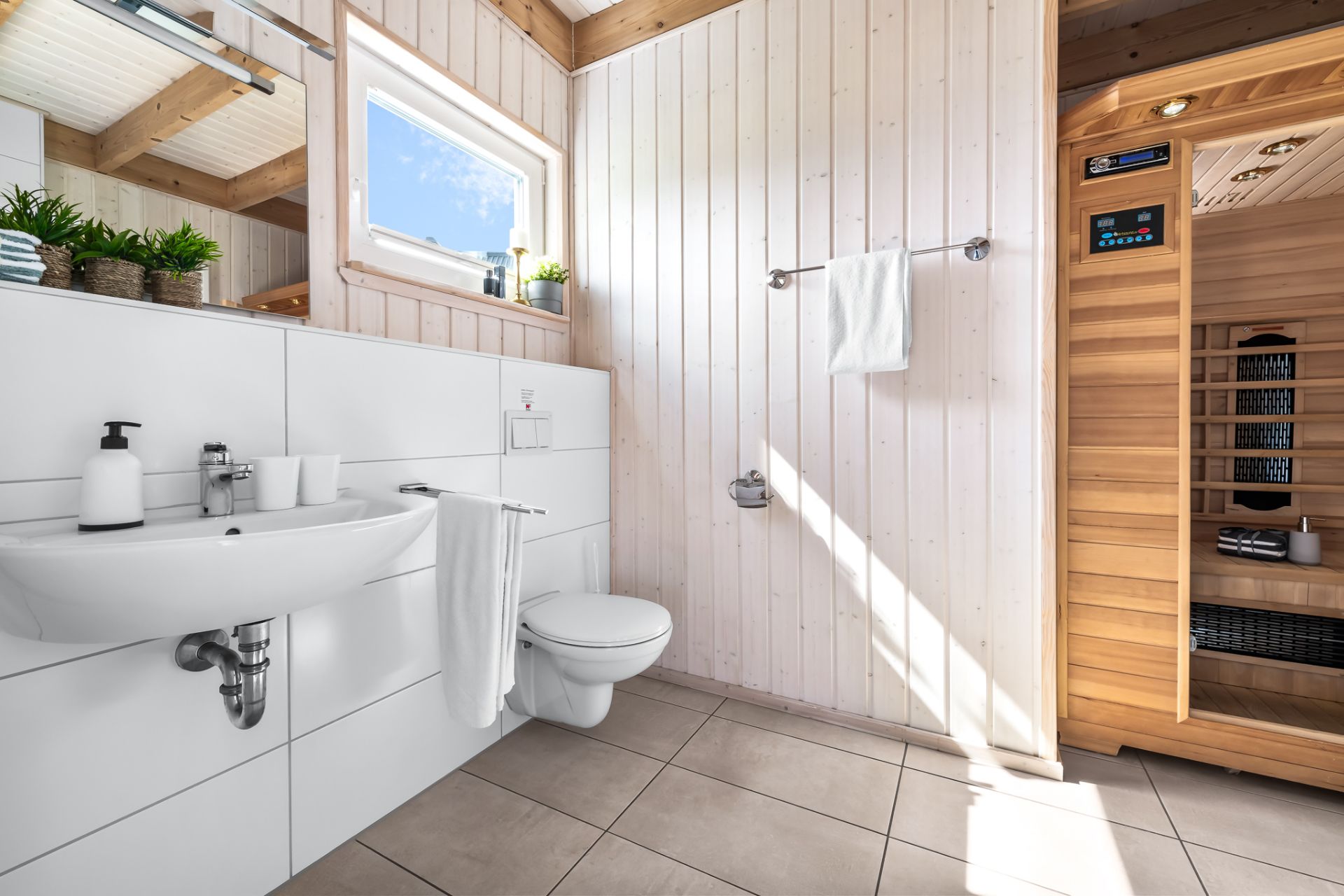 Helles Badezimmer mit Waschbecken, Wandtoilette und Zugang zu einer Holzsauna.