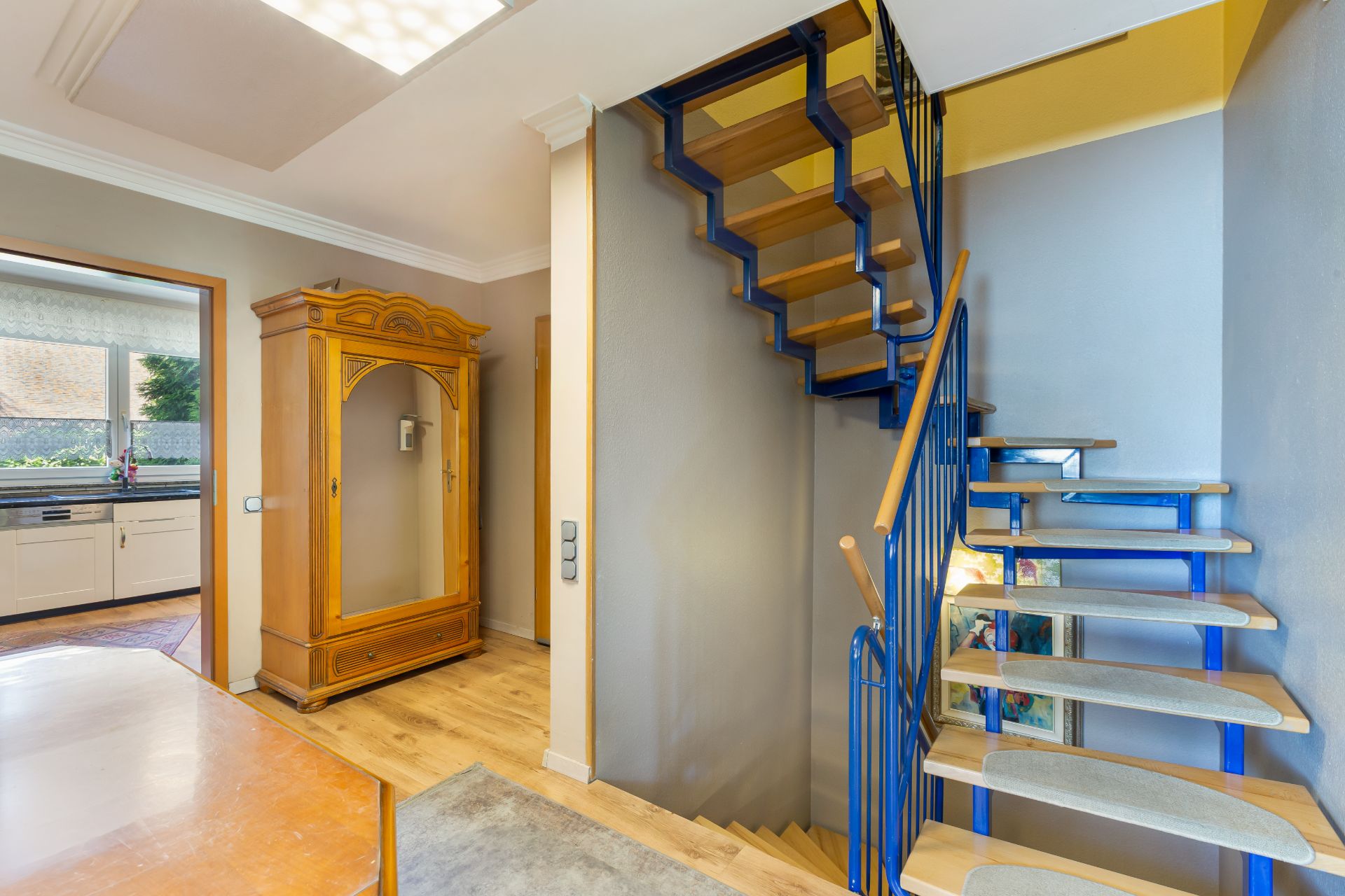 Innenraum mit moderner blauer Treppe, Holzschrank und Blick in eine helle Küche.