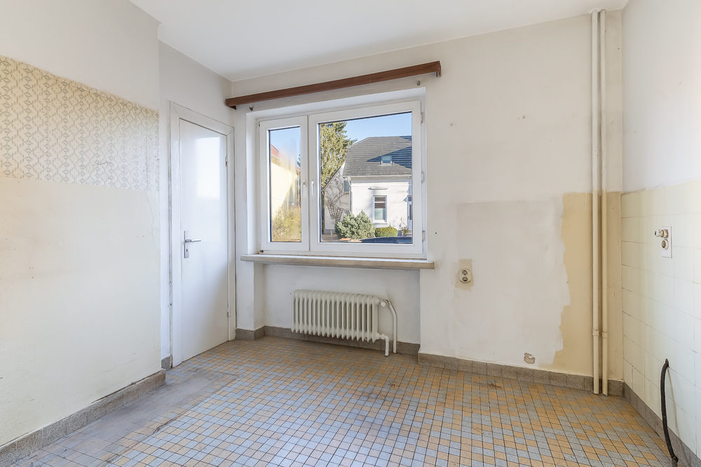 Leerer Raum mit Fliesenboden, weißer Tür, Fenster und Heizkörper unter dem Fenster.