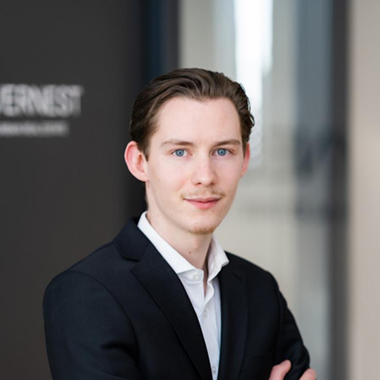 Junger Mann im Anzug steht mit verschränkten Armen in einem modernen Innenraum.